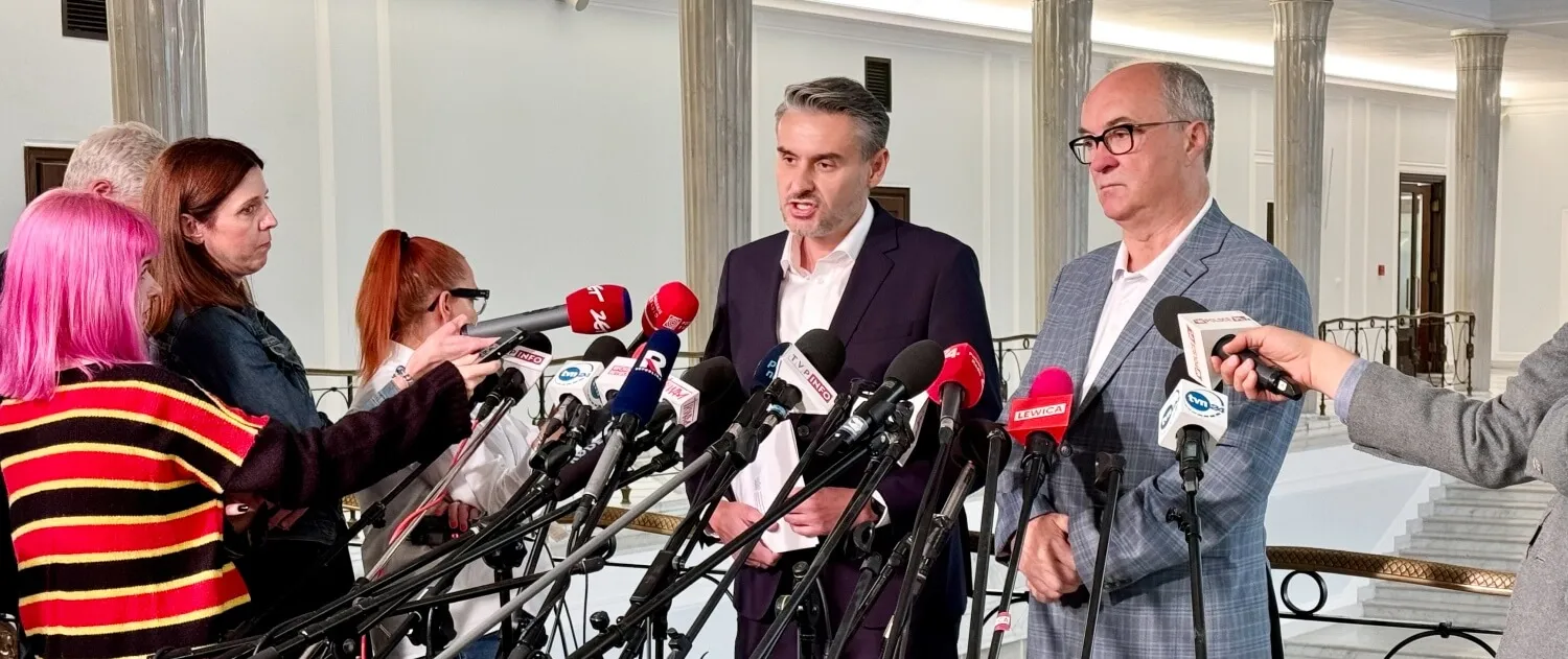 Mężczyzna w garniturze przemawia podczas konferencji prasowej Nowej Lewicy w sprawie zmian w Ministerstwie Sprawiedliwości, otoczony licznymi mikrofonami i dziennikarzami.
