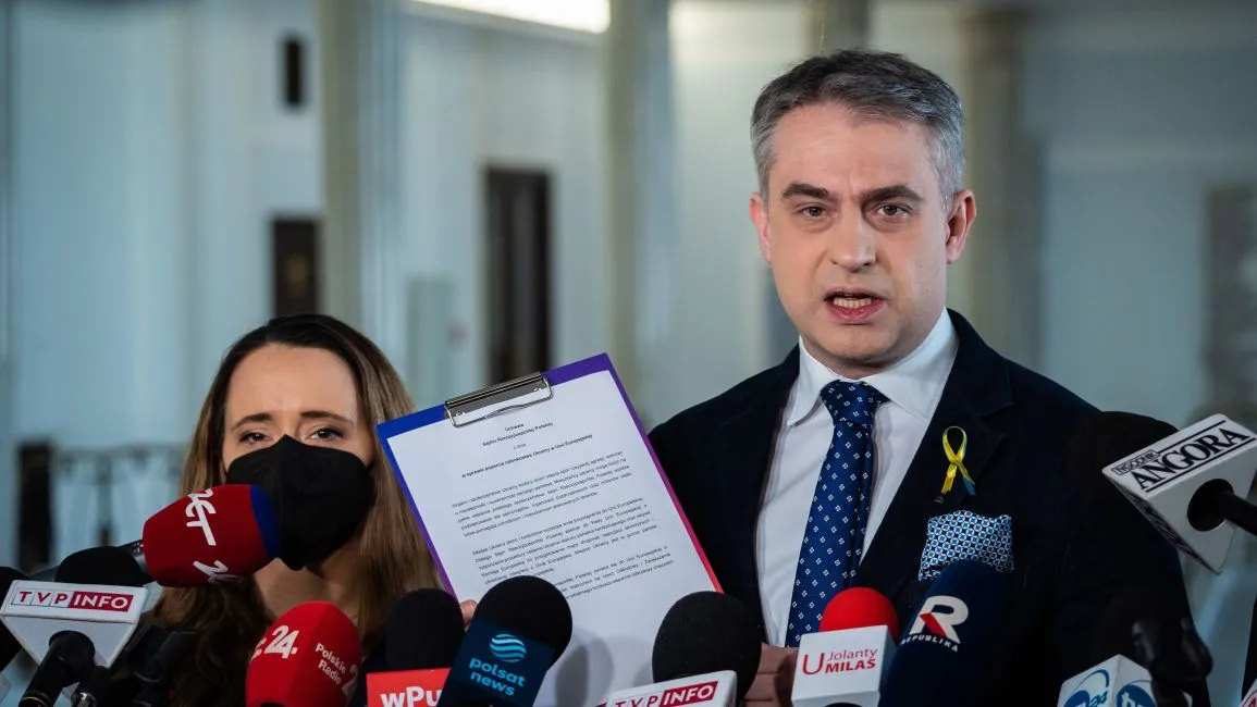 Mężczyzna w garniturze z żółtą wstążką na klapie trzyma na klipboardzie dokument podczas konferencji prasowej, otoczony przez liczne mikrofony mediów; obok niego stoi kobieta w maseczce.