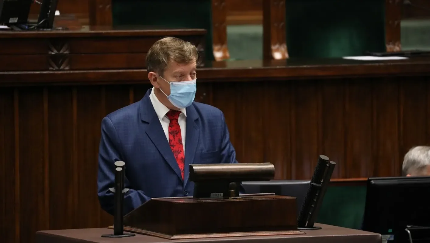 Polityk w garniturze i maseczce stoi przy mównicy w sali obrad.