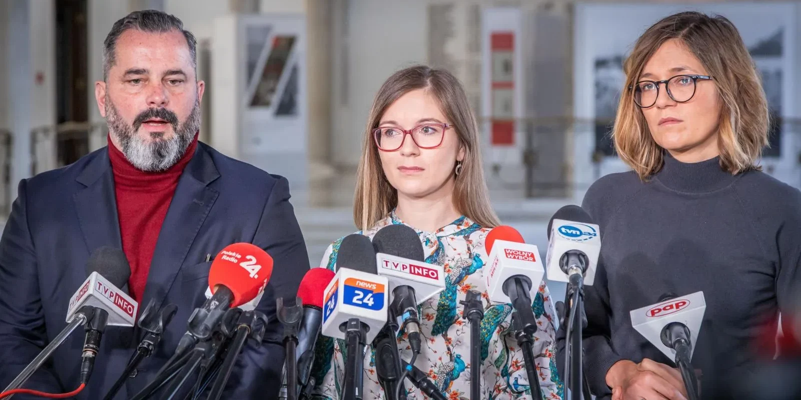 Na zdjęciu widoczni są trzej mówcy podczas konferencji prasowej, otoczeni mikrofonami i kamerami.