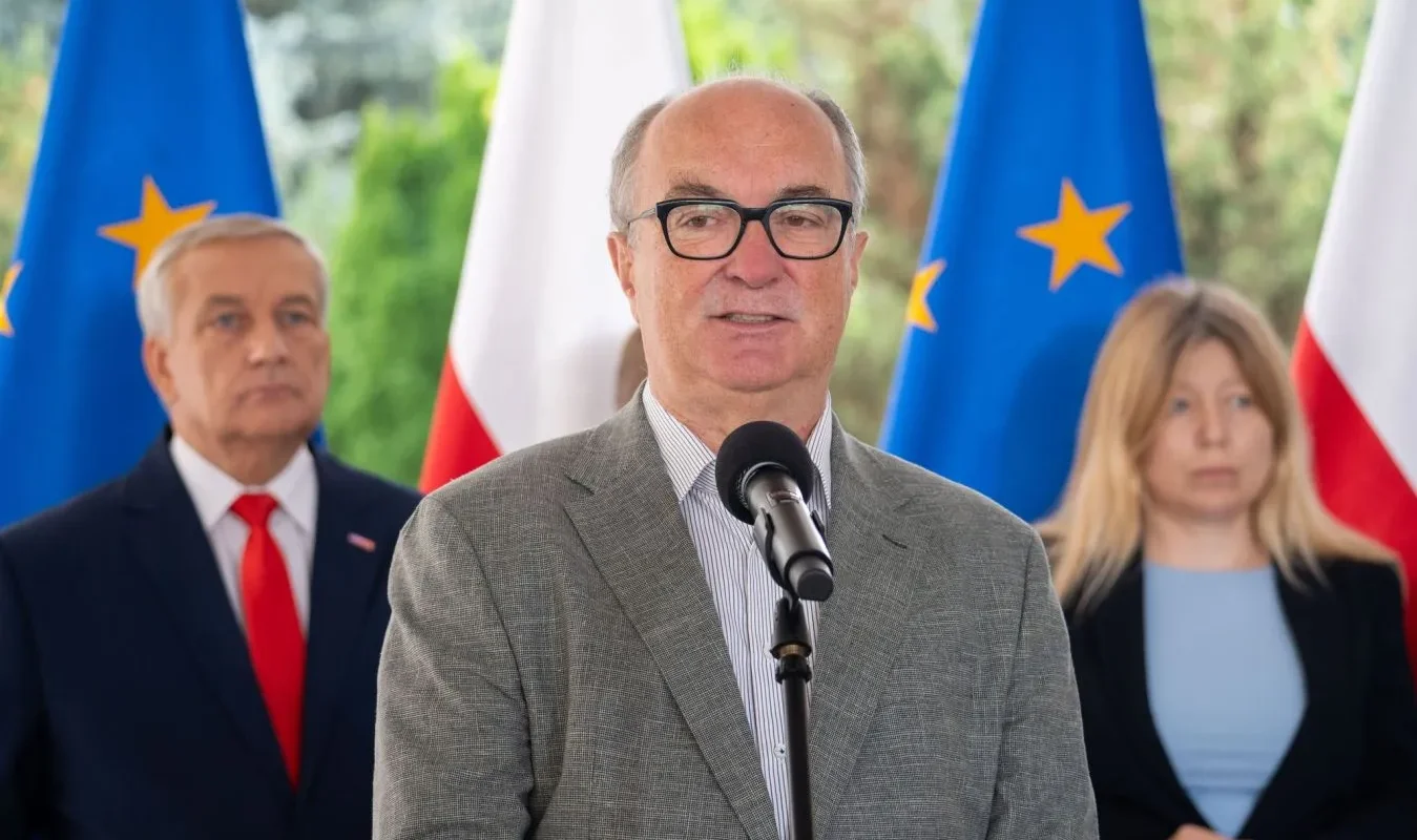 Mężczyzna w szarym garniturze i okularach przemawia do mikrofonu podczas konferencji prasowej, na tle flag UE i Polski.