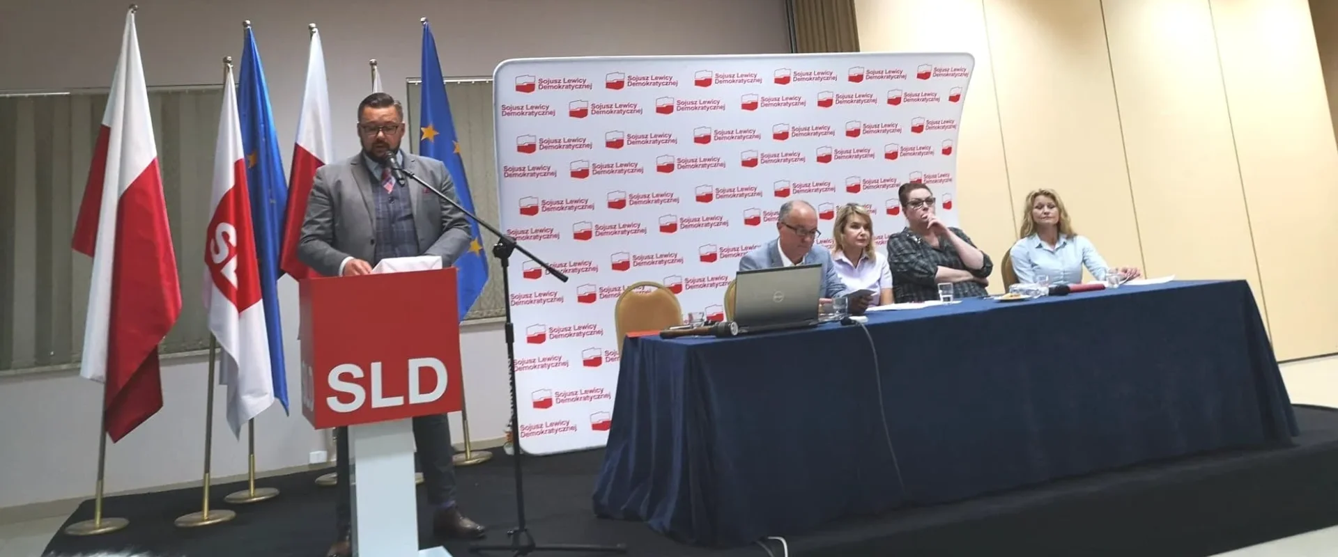 Mężczyzna w garniturze przemawia z podium podczas Rady Krajowej Sojuszu Lewicy Demokratycznej, na tle flag i panelu z kilkoma osobami siedzącymi przy stole.