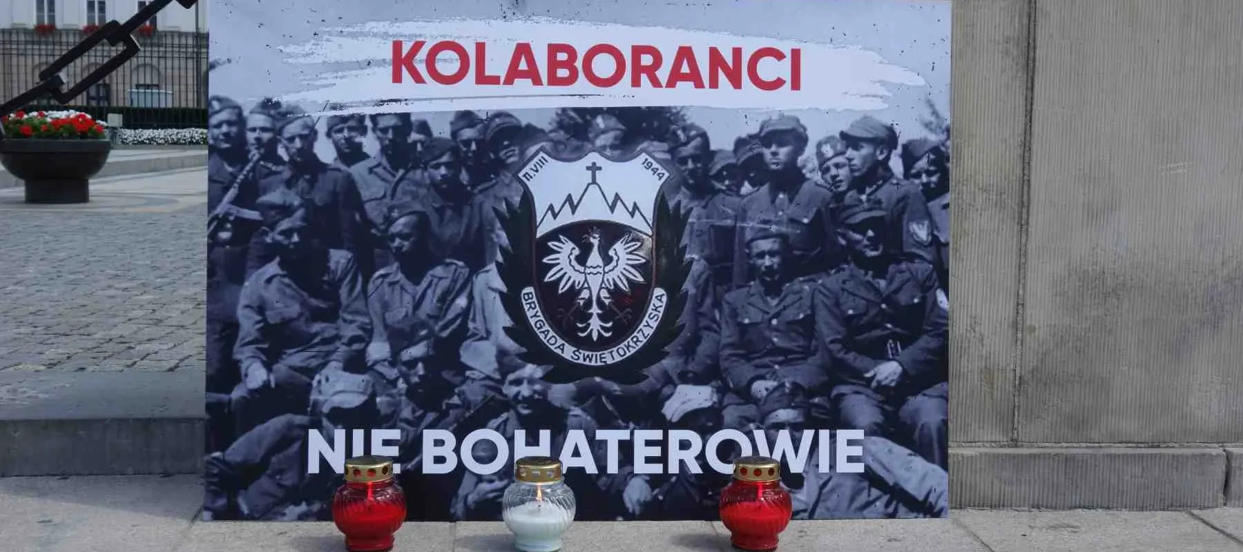 Duży czarno-biały plakat przedstawiający grupę żołnierzy z czerwonym napisem „KOLABORANCI” na górze i emblematem Brygady Świętokrzyskiej w środku oraz napisem „NIE BOHATEROWIE” na dole, stojący na placu.