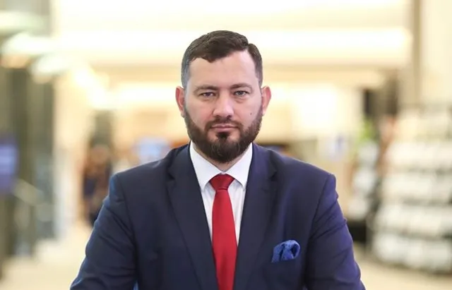 Mężczyzna z brodą w granatowym garniturze, czerwonym krawacie i niebieskiej poszetce stoi na tle jasnego, dobrze oświetlonego wnętrza.