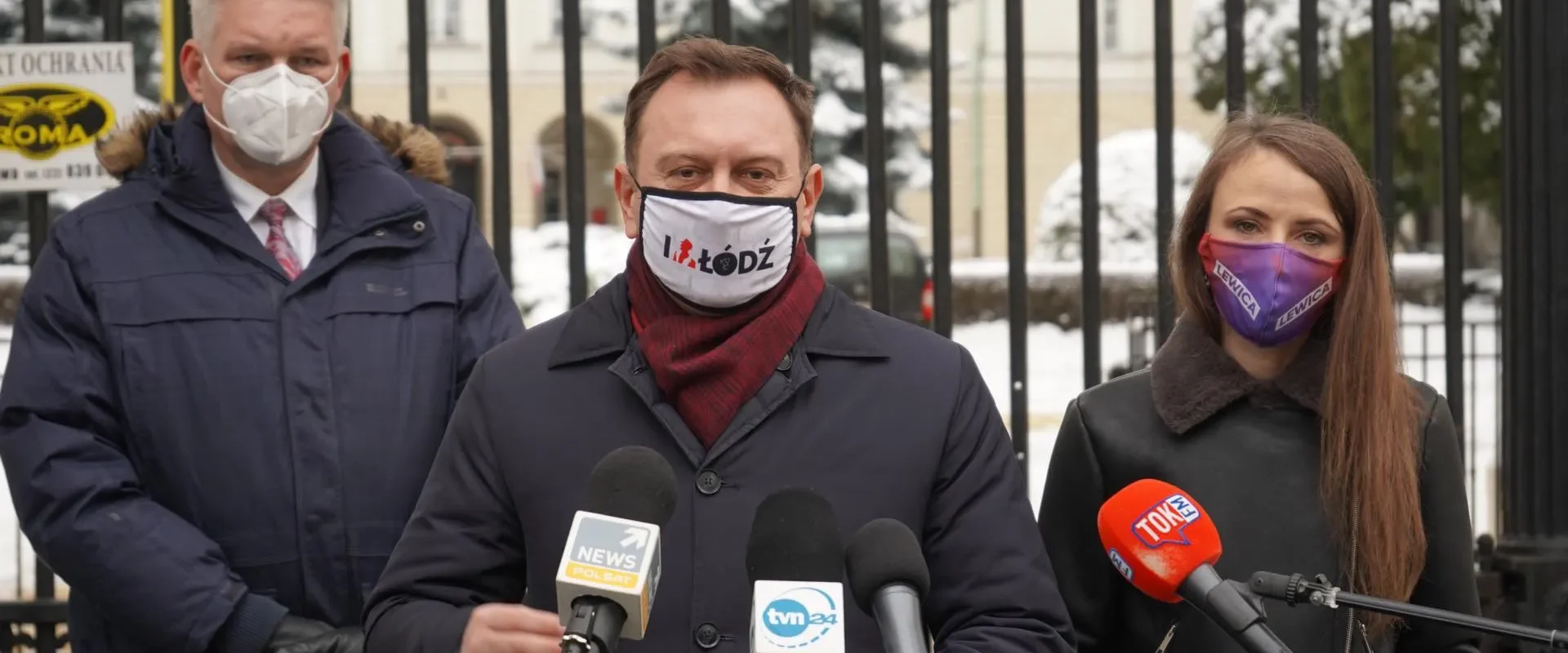 Trzy osoby w maseczkach stoją przed metalowym ogrodzeniem i przemawiają do mediów podczas konferencji prasowej na tle zimowego otoczenia.