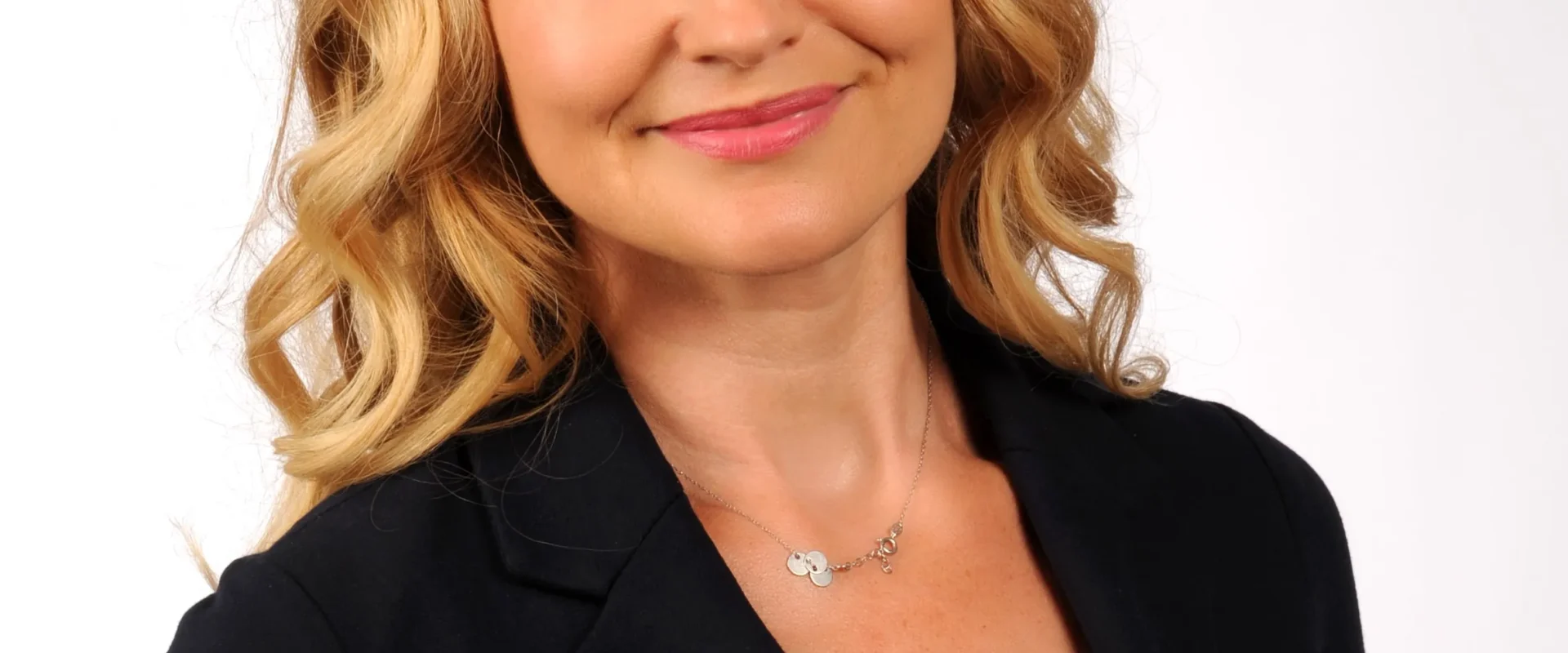 Portret uśmiechniętej kobiety o blond włosach, ubranej w czarny żakiet i białą bluzkę, na jasnym tle.