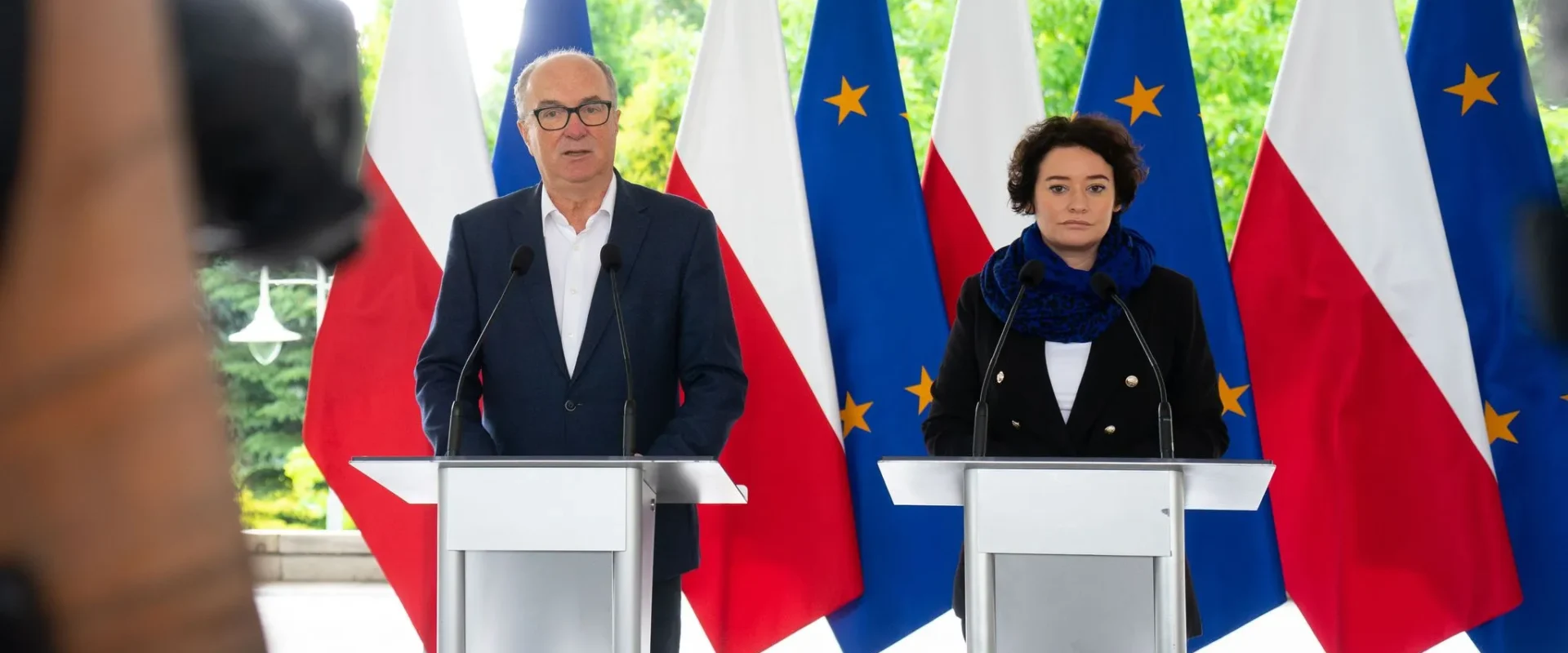 Dwóch polityków stoi przy oddzielnych podiumach z mikrofonami podczas konferencji prasowej, na tle flag Polski i Unii Europejskiej.