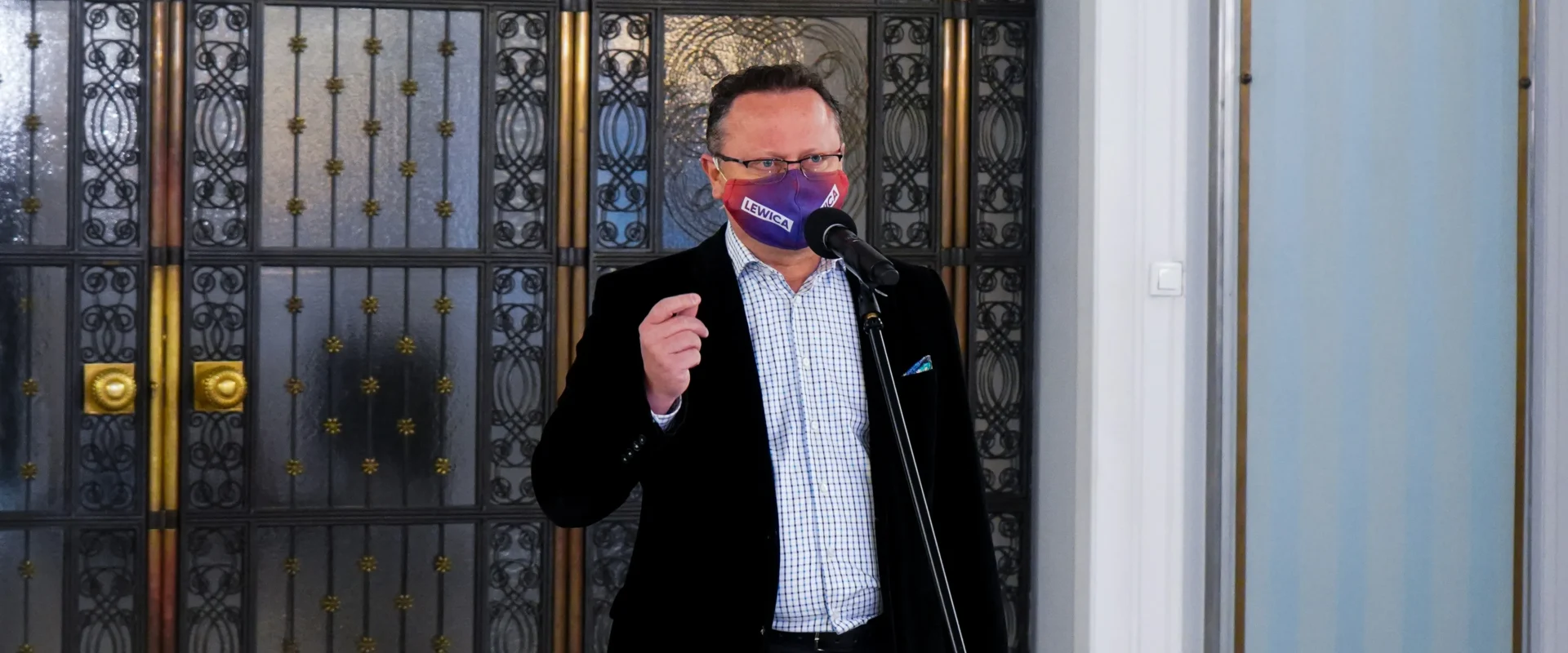 Mężczyzna w czarnej marynarce i koszuli stoi przy mikrofonie, nosi kolorową maseczkę z napisem LEWICA i stoi przed ozdobną metalową bramą.