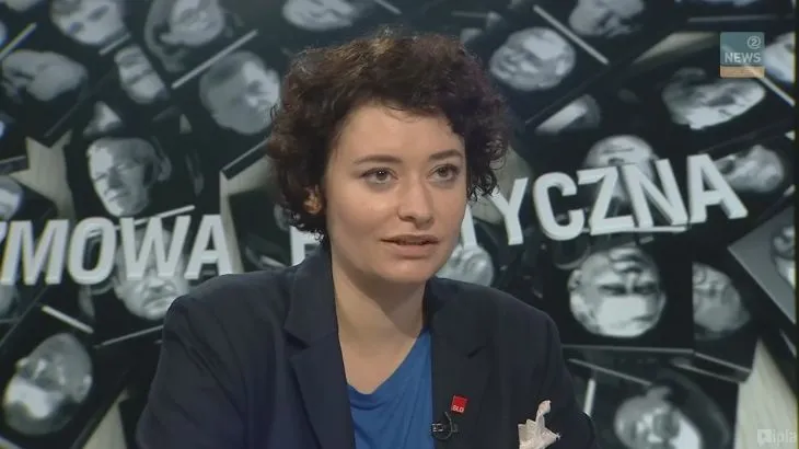 Kobieta w marynarce z krótkimi ciemnymi włosami udziela komentarza w studiu telewizyjnym na tle ekranu z napisami politycznymi, w programie o pozyskiwaniu środków z UE.