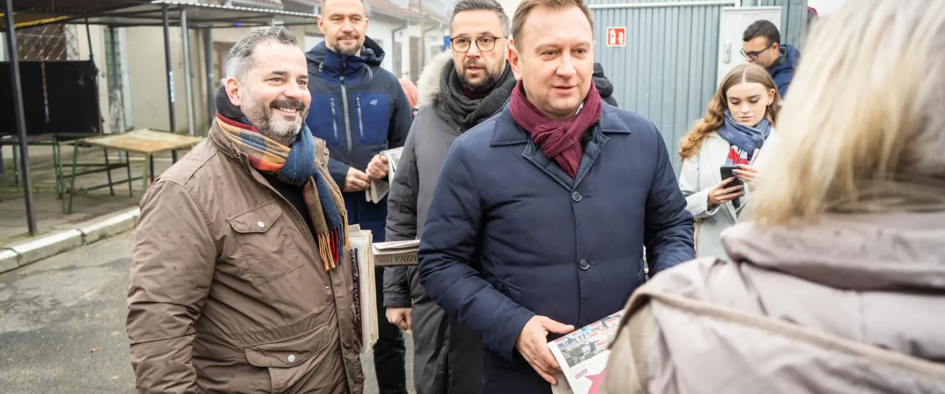 Na zdjęciu grupa mężczyzn i kobiet stoi na zewnątrz, rozmawiając i trzymając gazety, w otoczeniu naznaczonym ulicznym krajobrazem.
