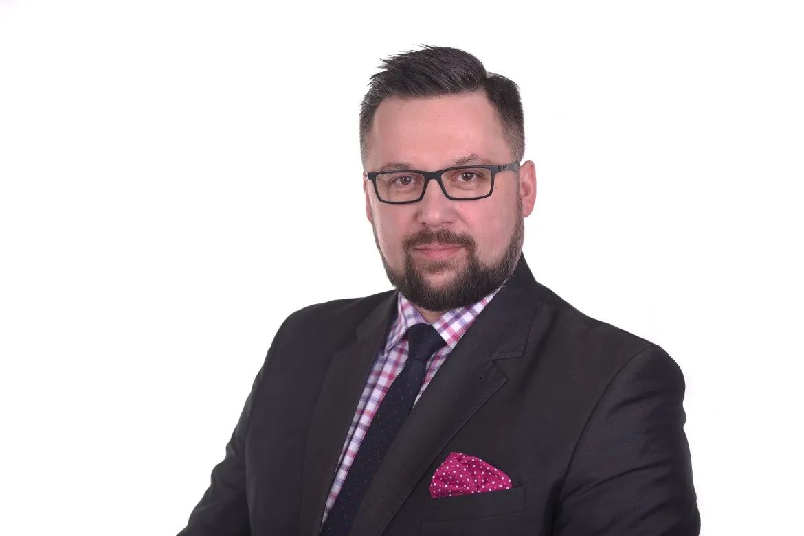 Portret mężczyzny w czarnym garniturze z różową poszetką i okularami na białym tle.