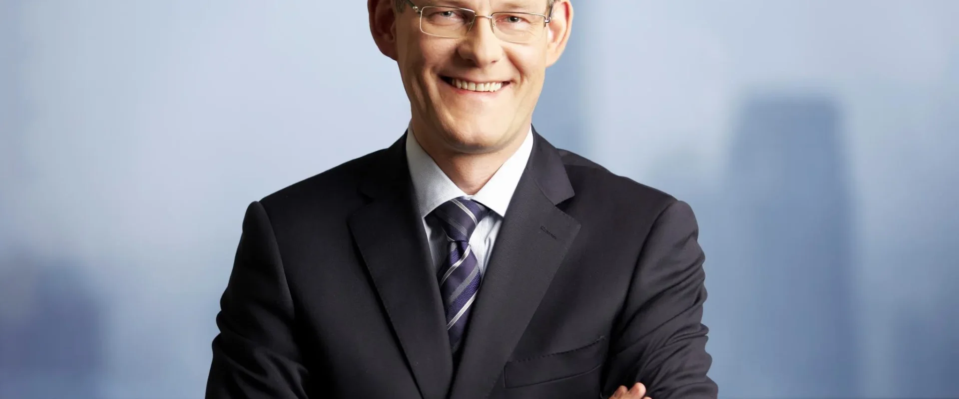 Uśmiechnięty mężczyzna w garniturze stoi na tle rozmytego widoku miasta, z założonymi rękami.