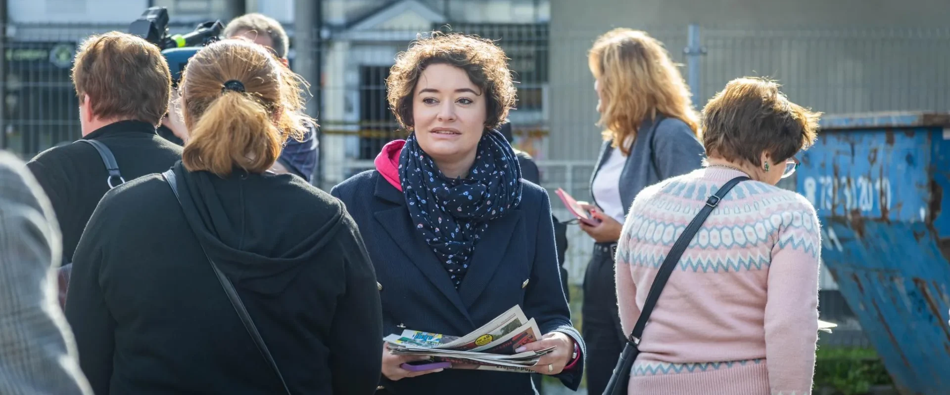 Kobieta w granatowej marynarce i szaliku trzyma gazety, stojąc wśród grupy ludzi na zewnątrz w miejskim otoczeniu.