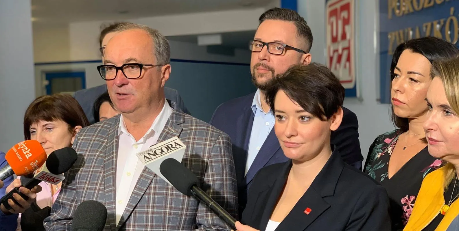Grupa polityków lewicy podczas konferencji prasowej; mężczyzna w marynarce w kratkę przemawia do reporterów, otoczony innymi osobami, z mikrofonami mediów w tle.