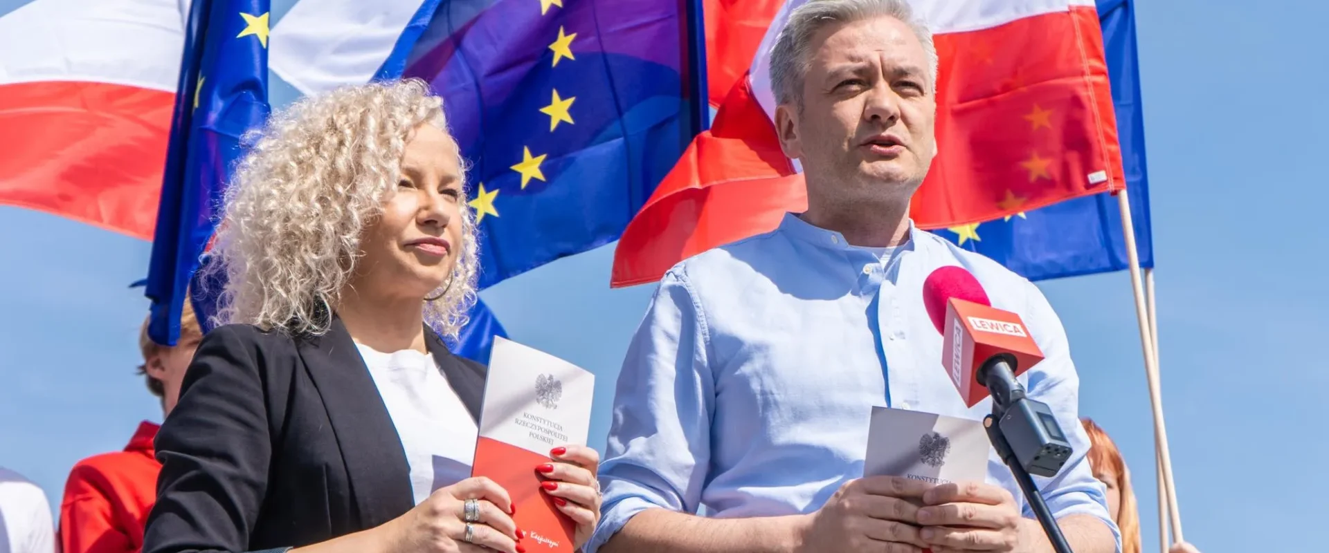 Dwóch mówców na wydarzeniu politycznym w Polsce: mężczyzna w jasnoniebieskiej koszuli przemawia do mikrofonu, trzymając kartkę z godłem, a obok niego stoi kobieta z broszurą; w tle widoczne flagi Polski i Unii Europejskiej.