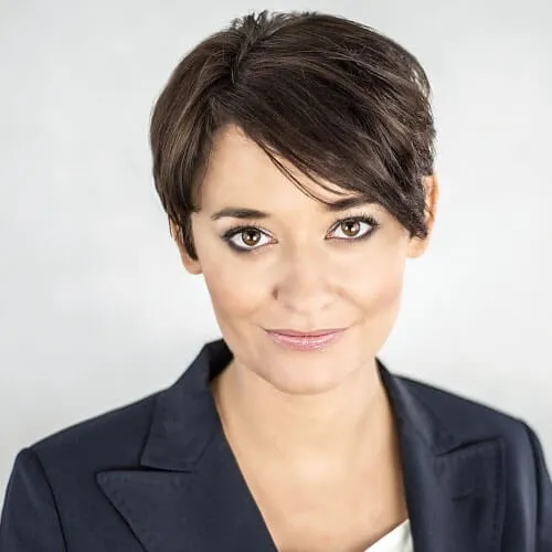 Portret kobiety o krótkich ciemnych włosach, uśmiechniętej, w ciemnym garniturze na jasnym tle.