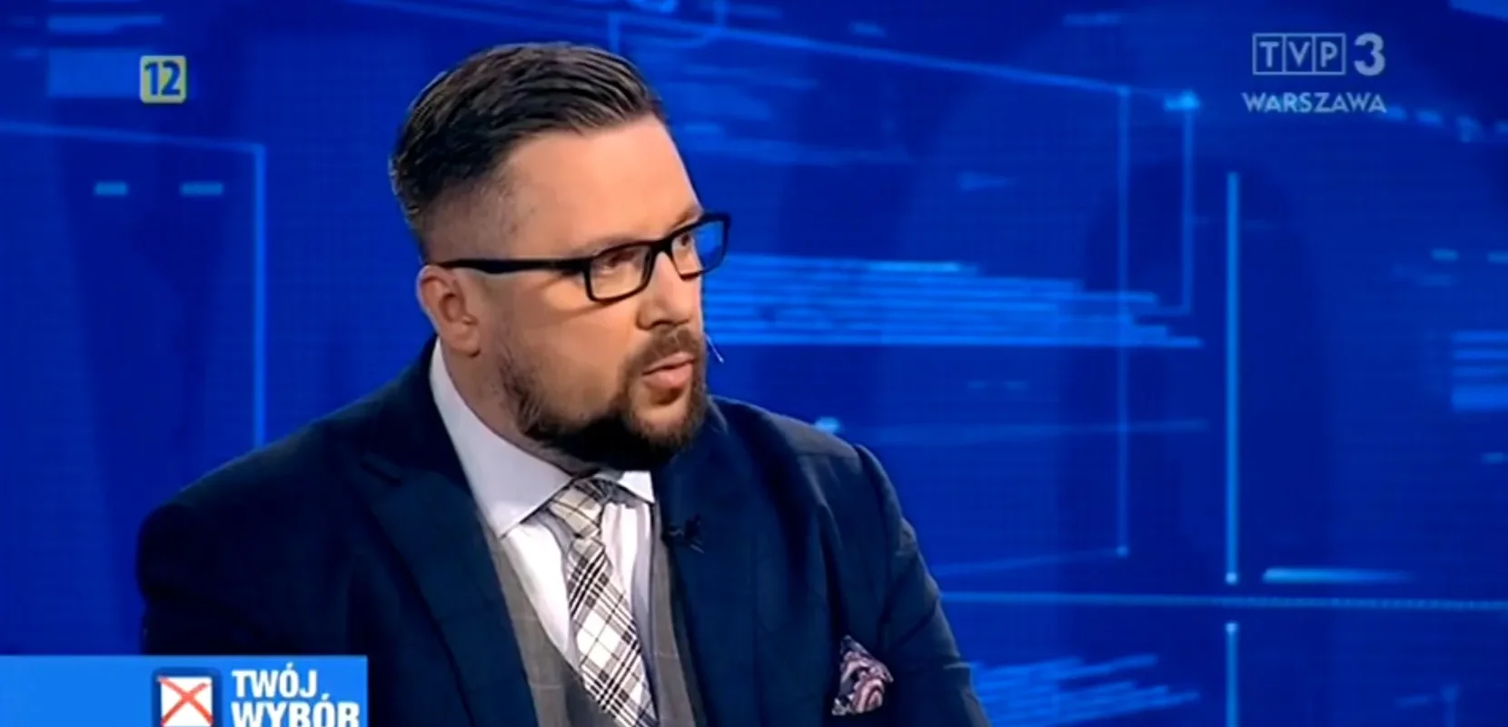 Mężczyzna w garniturze i okularach rozmawia w studiu telewizyjnym TVP3 Warszawa, na tle niebieskiej grafiki z logotypem stacji i napisem „Twój Wybór”.
