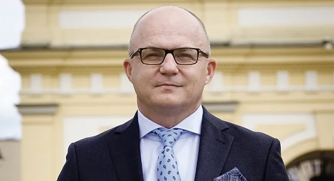 Portret mężczyzny w garniturze i okularach na tle żółtej fasady budynku.