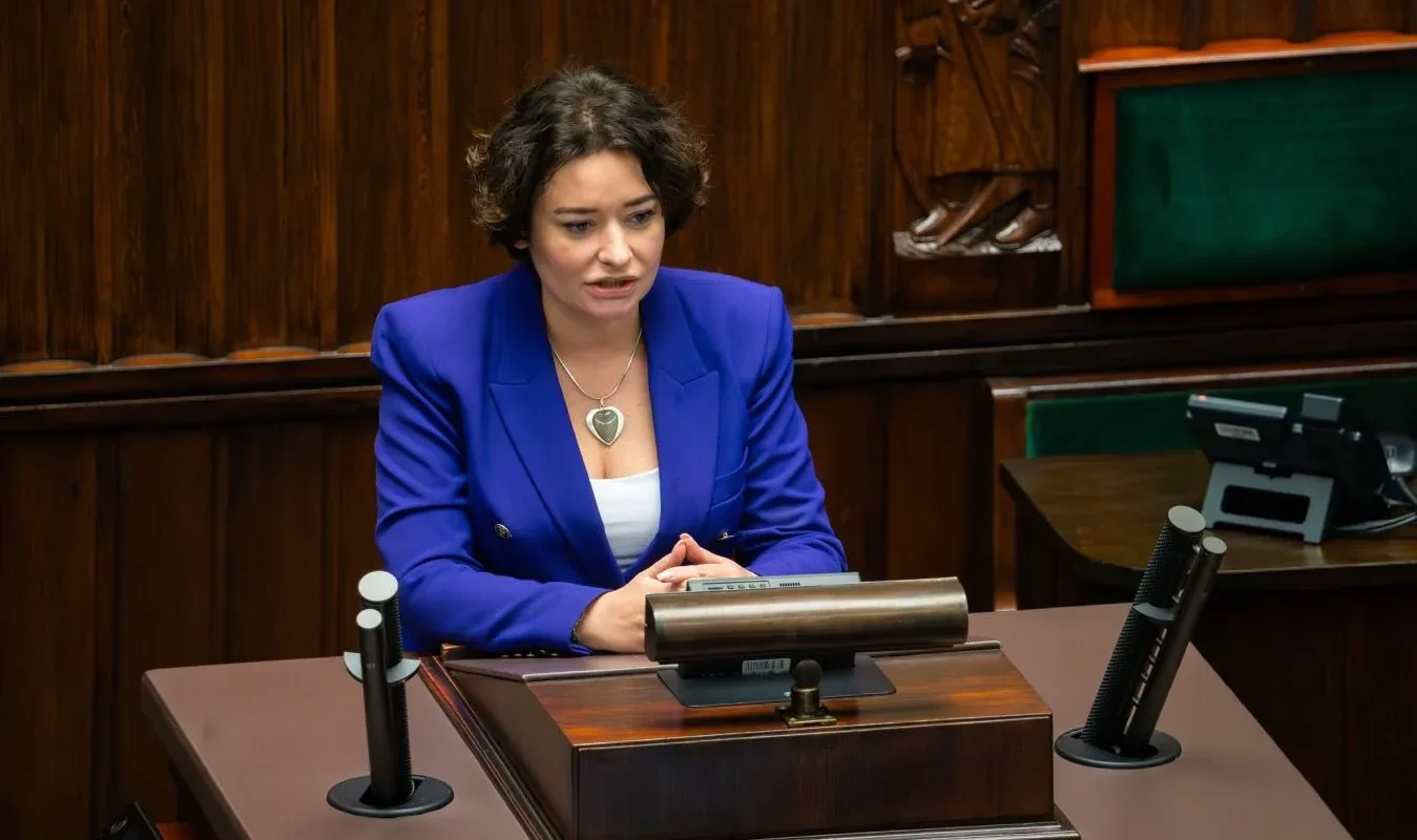 Kobieta w niebieskiej marynarce siedzi za pulpitem w sali obrad, otoczona mikrofonami i drewnianymi panelami.