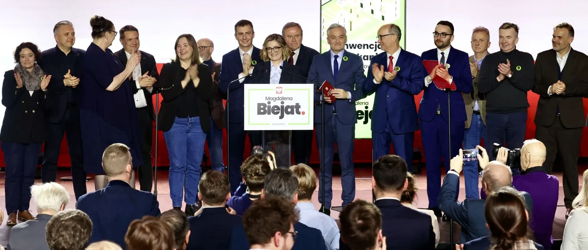 Grupa osób stojących na scenie podczas konferencji politycznej, wokół mównicy z plakatem kampanii, a przed sceną publiczność oklaskuje.