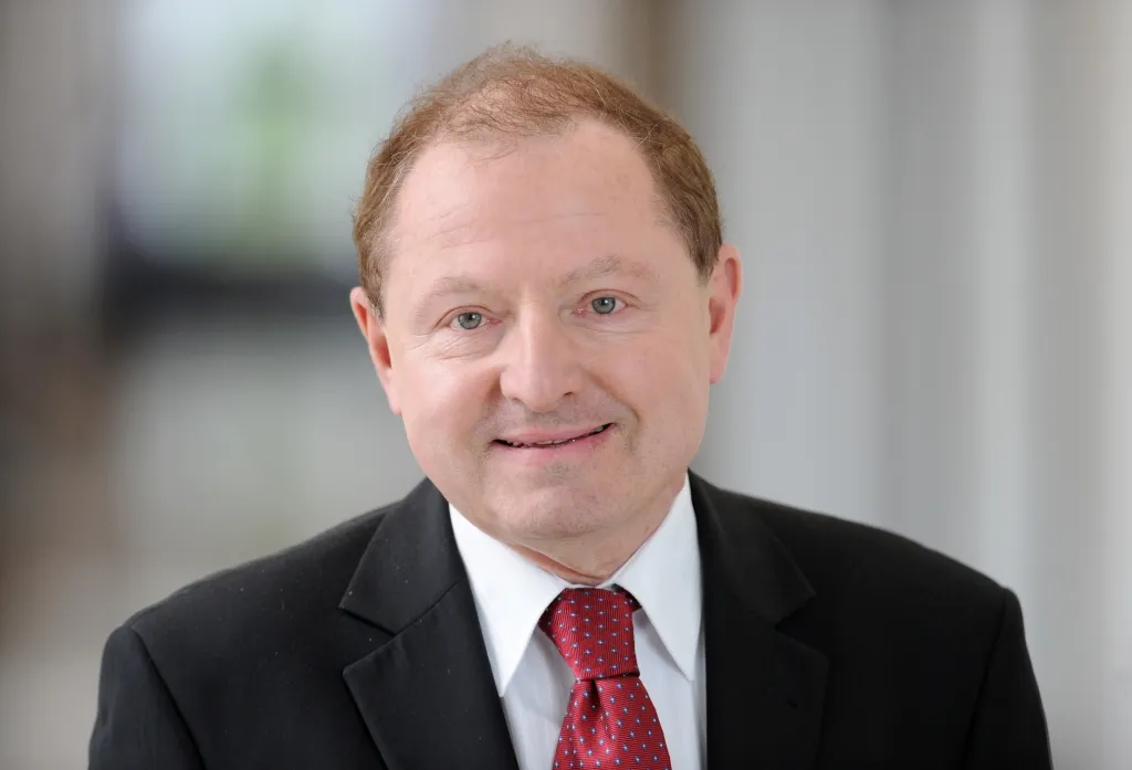 Portret mężczyzny w czarnym garniturze i czerwonym krawacie, uśmiechnięty, na neutralnym, rozmytym tle.