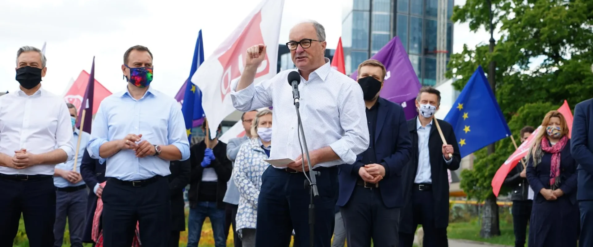 Mężczyzna z okularami wygłasza przemówienie do mikrofonu podczas plenerowej konferencji, otoczony przez innych uczestników w maseczkach, z flagami w tle i widocznym nowoczesnym budynkiem.