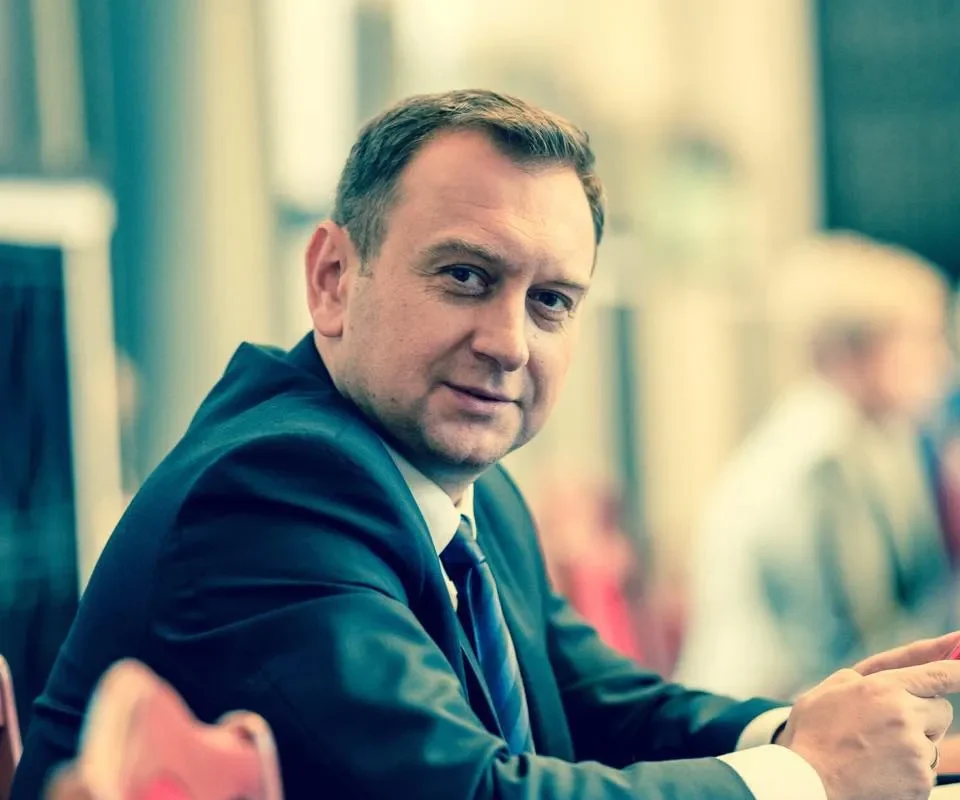 Mężczyzna w garniturze siedzi przy stole i patrzy w kamerę na tle rozmytego wnętrza.