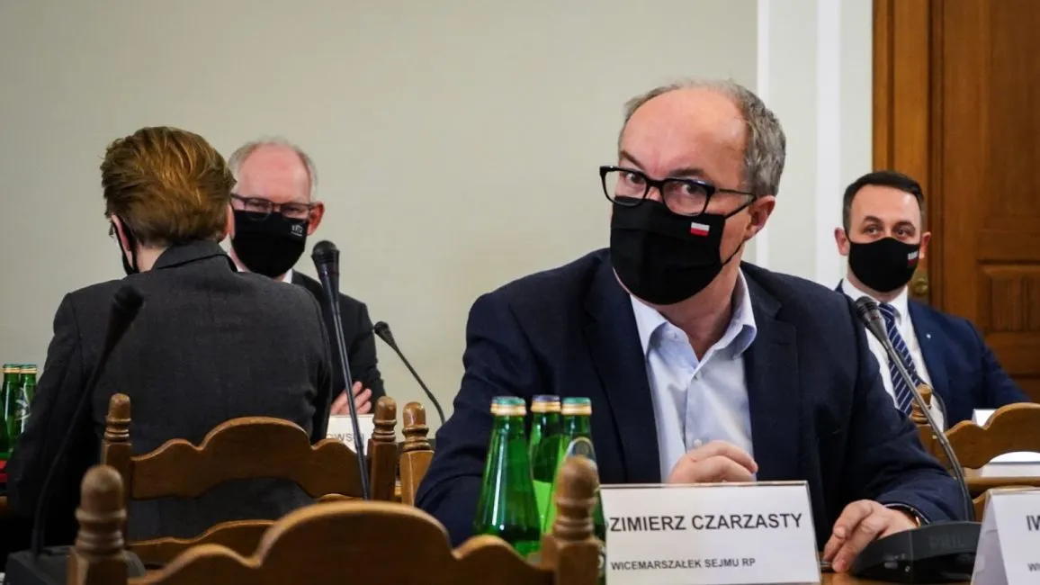 Grupa osób w garniturach podczas posiedzenia; na pierwszym planie mężczyzna w okularach i czarnej maseczce siedzi przy stole z mikrofonem, obok widoczne zielone butelki.