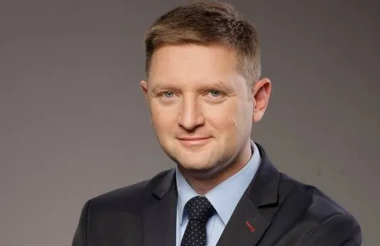 Portret mężczyzny w czarnym garniturze, białej koszuli i granatowym krawacie na neutralnym tle, z lekkim uśmiechem.