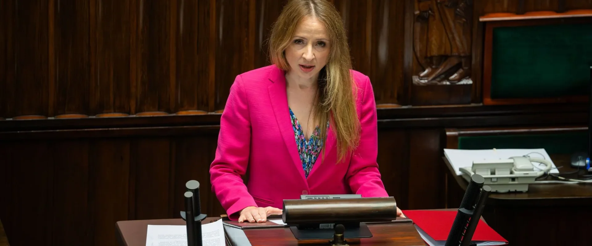 Kobieta w różowej marynarce stoi przy mównicy w sali obrad, wygłasza przemówienie.