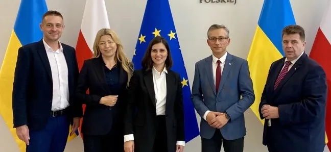Na zdjęciu stoi pięć osób w formalnych ubraniach, z flagami Polski, Ukrainy oraz Unii Europejskiej w tle.
