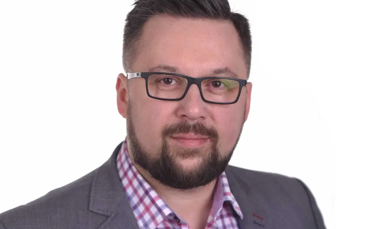 Portret mężczyzny w okularach, w szarym garniturze i koszuli w kratę na białym tle.