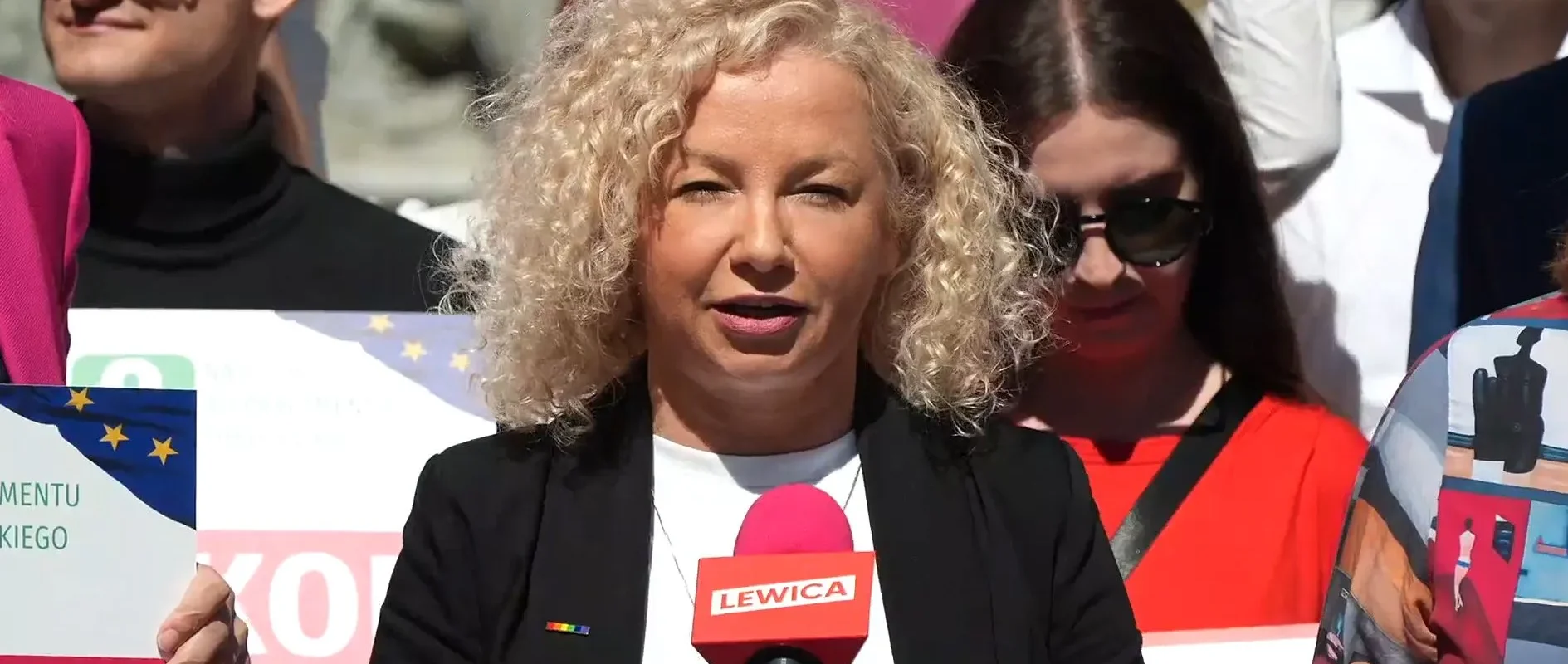 Kobieta o kręconych blond włosach przemawia do mikrofonu z napisem LEWICA podczas wiecu, wokół stoją uczestnicy z plakatami.