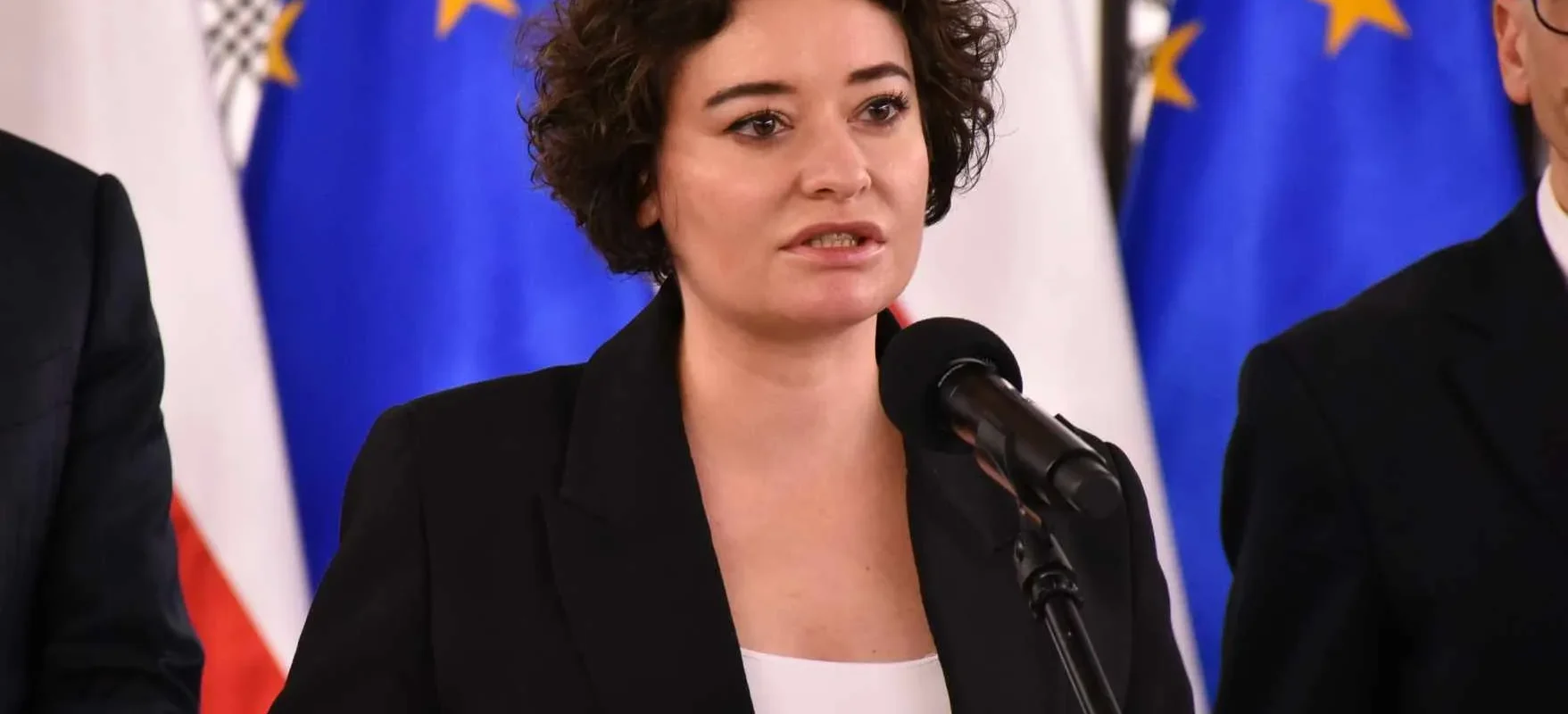 Kobieta w czarnym garniturze przemawia do mikrofonu podczas konferencji, na tle flag Unii Europejskiej.