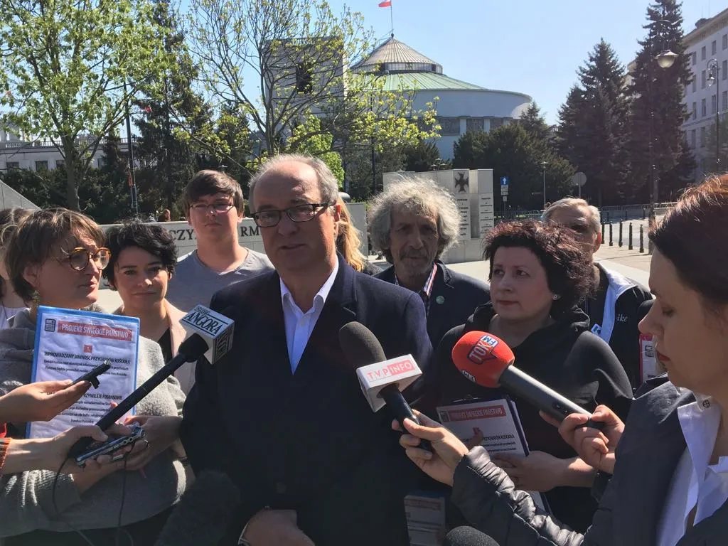 Mężczyzna w garniturze udziela wywiadu przed kamerami i mikrofonami dziennikarzy podczas konferencji prasowej na placu z budynkami w tle.
