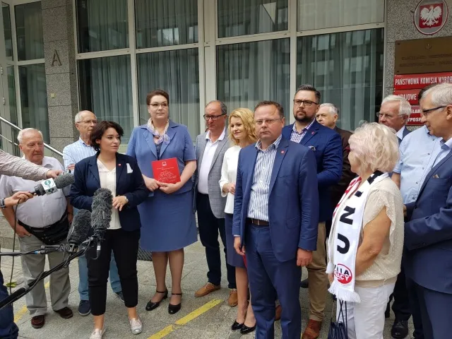 Grupa osób w formalnych ubraniach stojąca na zewnątrz budynku przy mikrofonach, podczas konferencji prasowej związanej z rejestracją Komitetu SLD Lewica Razem.