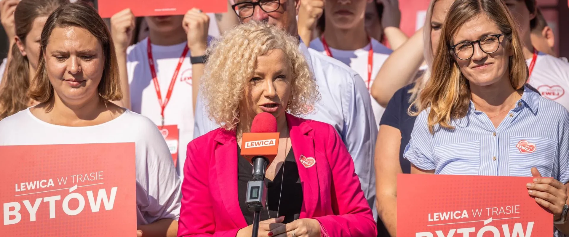 Grupa działaczy Lewicy na wiecu w Bytowie, na pierwszym planie kobieta w różowej marynarce mówiąca do mikrofonu, trzymająca plakat z napisem Bytów.