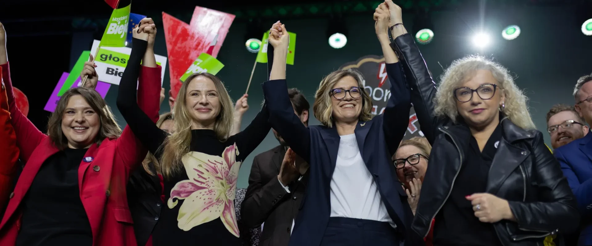 Grupa kobiet na scenie podczas wiecu wyborczego, uniesionymi rękami w geście zwycięstwa, w tle kolorowe plakaty i flaga Polski.