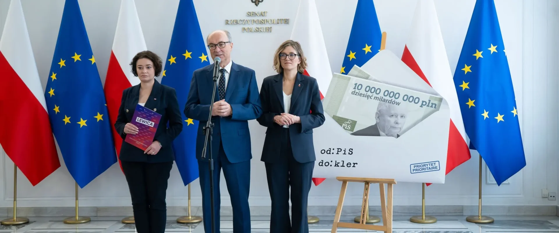 Trzy osoby w garniturach stoją podczas konferencji, na tle flag Polski i Unii Europejskiej, obok dużej makiety banknotu z napisem „10 000 000 000 zł” i hasłem „od PiS do: kler”.