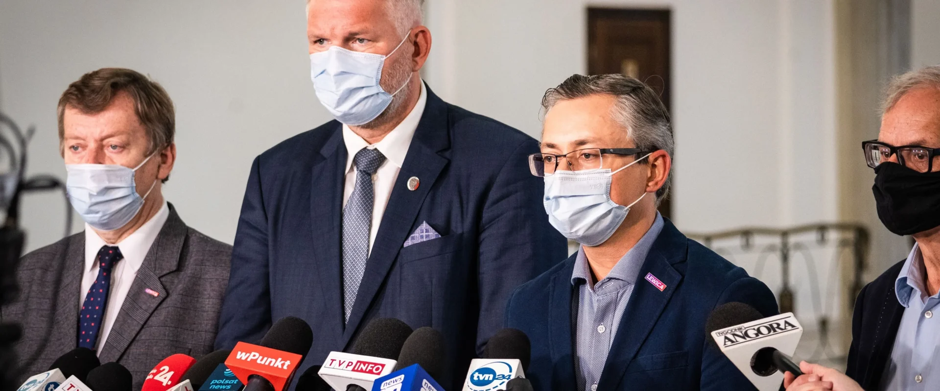 Grupa mężczyzn w garniturach i maseczkach stoi na konferencji prasowej, otoczona licznymi mikrofonami dziennikarzy.
