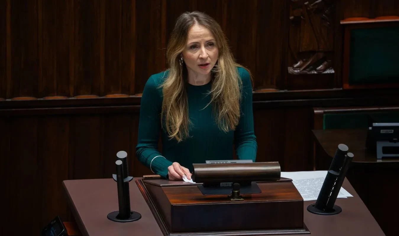 Kobieta o długich blond włosach w zielonej bluzce stoi przy mównicy w sali obrad, otoczona drewnianym wystrojem i mikrofonami.