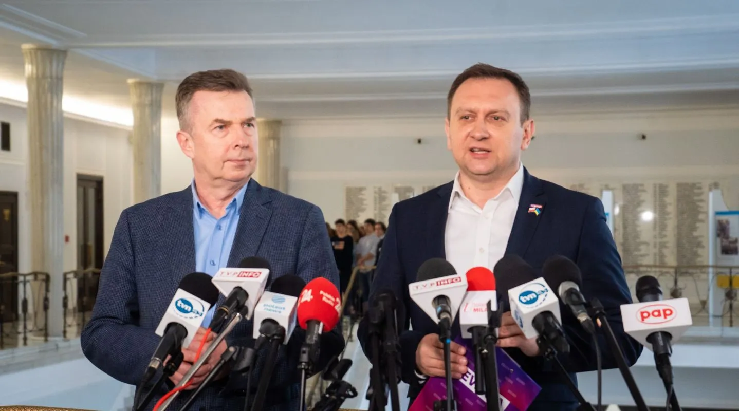 Dwaj mężczyźni w garniturach stoją przy mównicy z mikrofonami, rozmawiając podczas konferencji prasowej w stylowym wnętrzu.