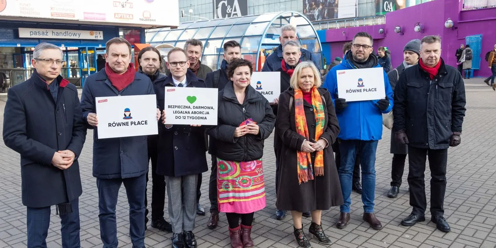 Grupa osób trzymających plakaty z hasłami wzywającymi do równych płac oraz legalnej aborcji, stojąca przed pasażem handlowym.