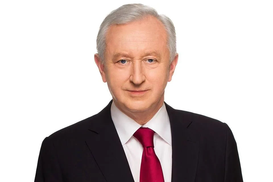Portret starszego mężczyzny w ciemnym garniturze i bordowym krawacie na białym tle, patrzącego prosto w obiektyw.