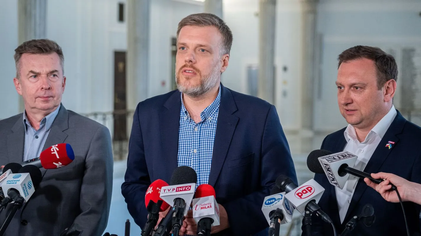 Na zdjęciu trzej mężczyźni stoją przed mikrofonami, prowadząc konferencję prasową w przestronnej, jasnej sali.