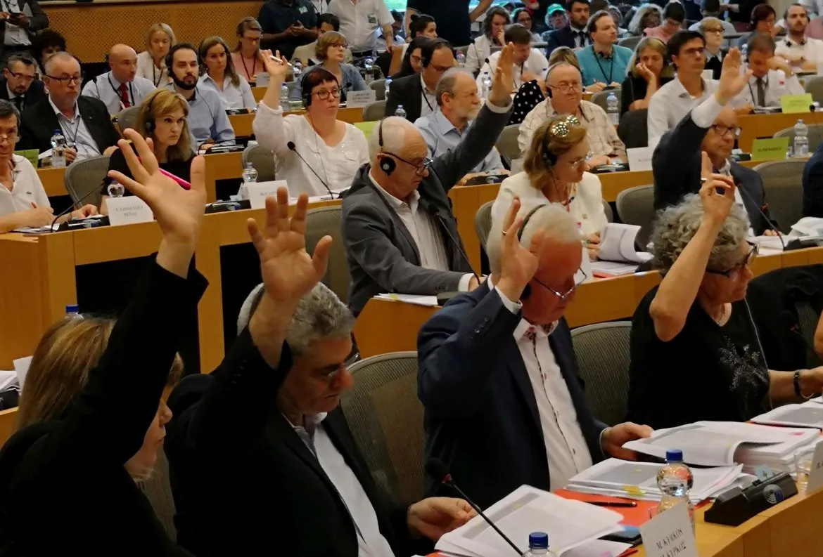Grupa uczestników konferencji dla polskich przewoźników podnosi ręce podczas sesji, siedząc przy stołach z dokumentami i mikrofonami.