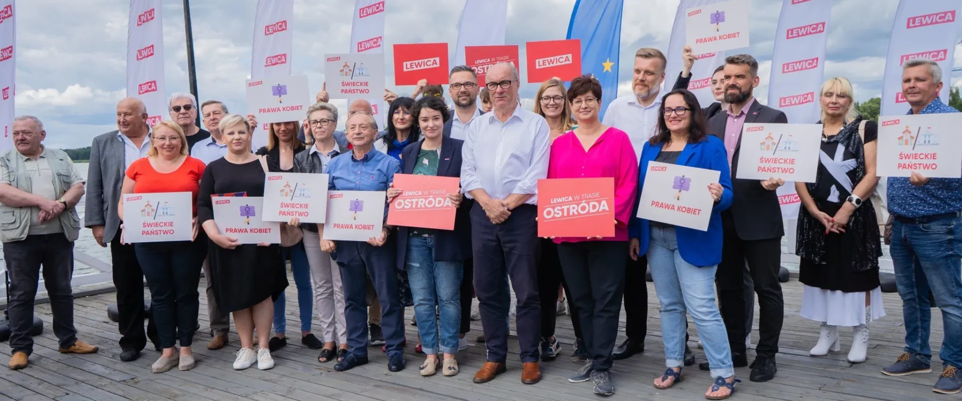 Grupa uczestników marszu 1 października stoi na drewnianym pomoście nad wodą, trzymając plakaty z hasłami Lewica, Prawa Kobiet i Świeckie państwo.