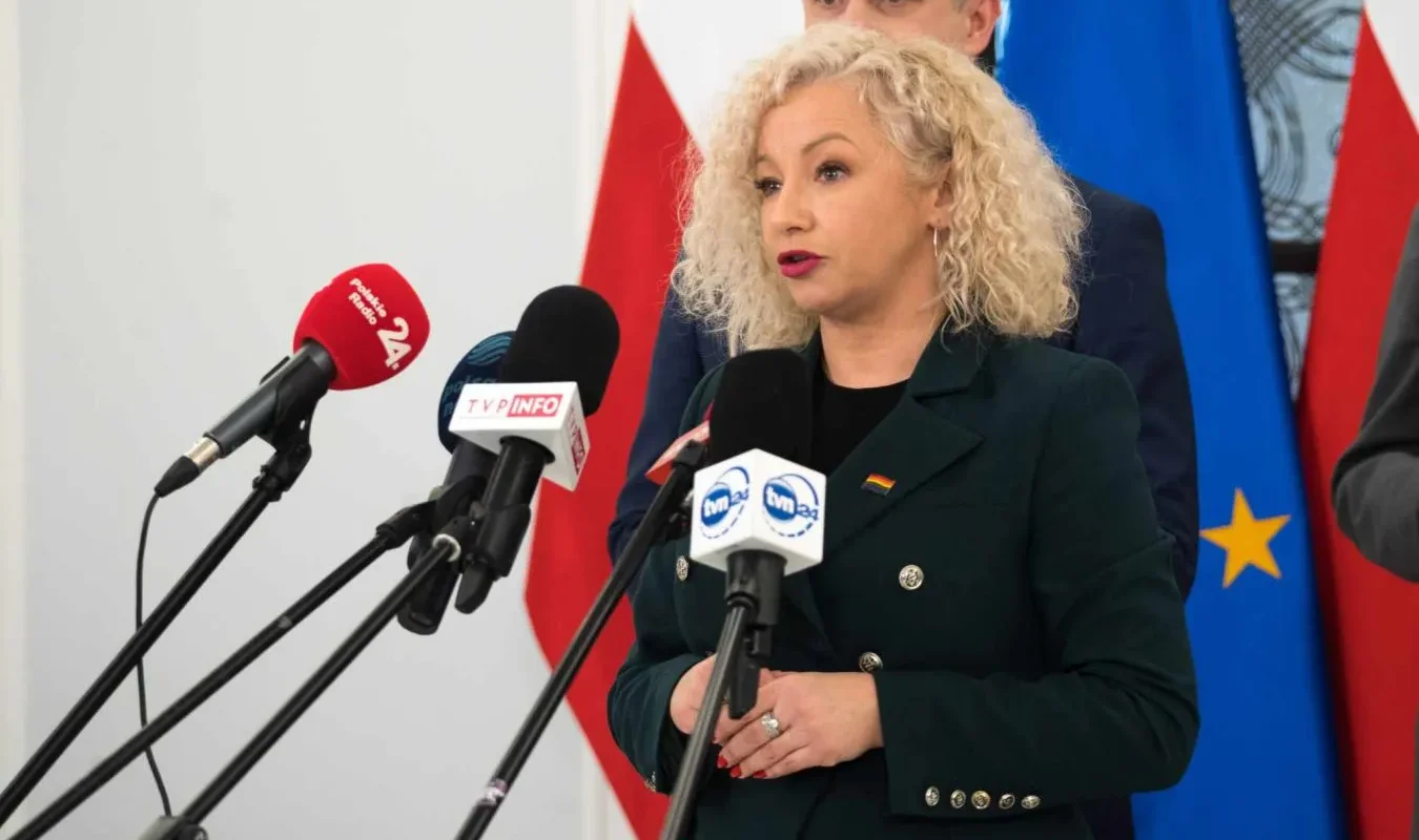 Polityczka w zielonej marynarce przemawia na konferencji prasowej, stojąc przy mikrofonach, z flagami państwowymi w tle.