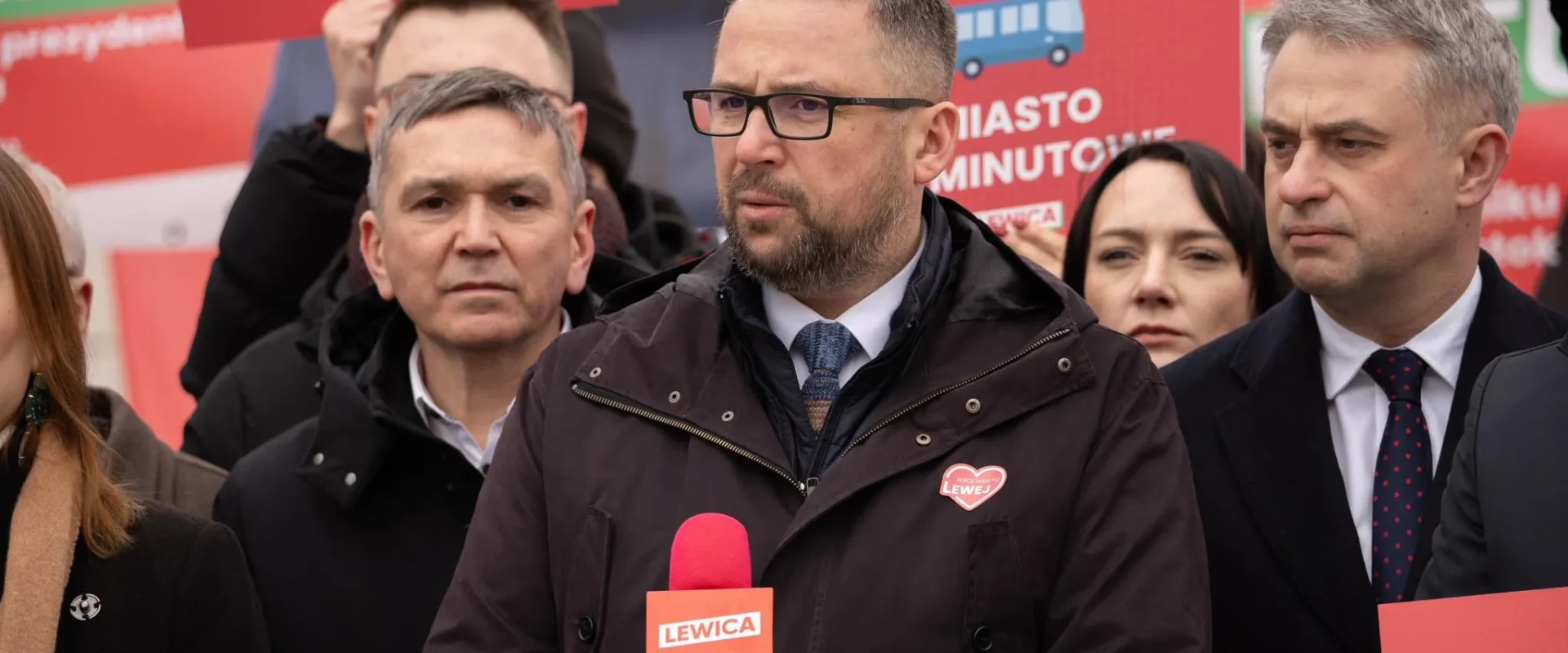 Polityk w brązowym płaszczu i okularach przemawia do mikrofonu Lewica podczas wiecu wyborczego, otoczony innymi kandydatami i czerwonymi plakatami w tle.