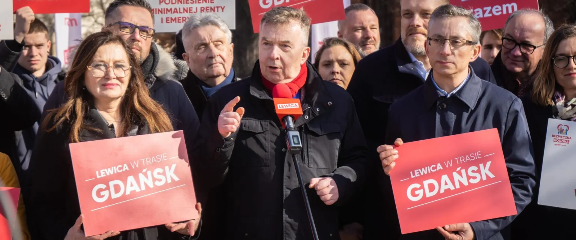 Grupa zwolenników Lewicy podczas wiecu, mężczyzna przemawia do mikrofonu na tle czerwonych plakatów z napisami LEWICA i GDANSK.
